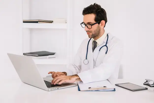 Tired doctor working late at his desk surrounded by paperwork, showing the burden of documentation without a virtual medical scribe.
